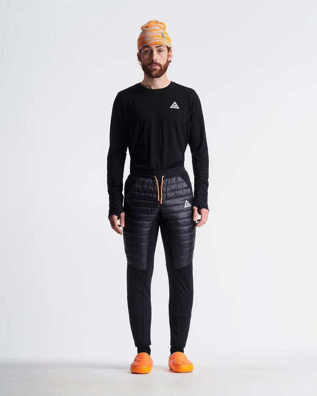 A man stands against a light gray studio background wearing a black long-sleeve athletic top with a small white logo on the chest, black quilted pants with a gold drawstring, a yellow and orange striped beanie, and bright orange shoes.