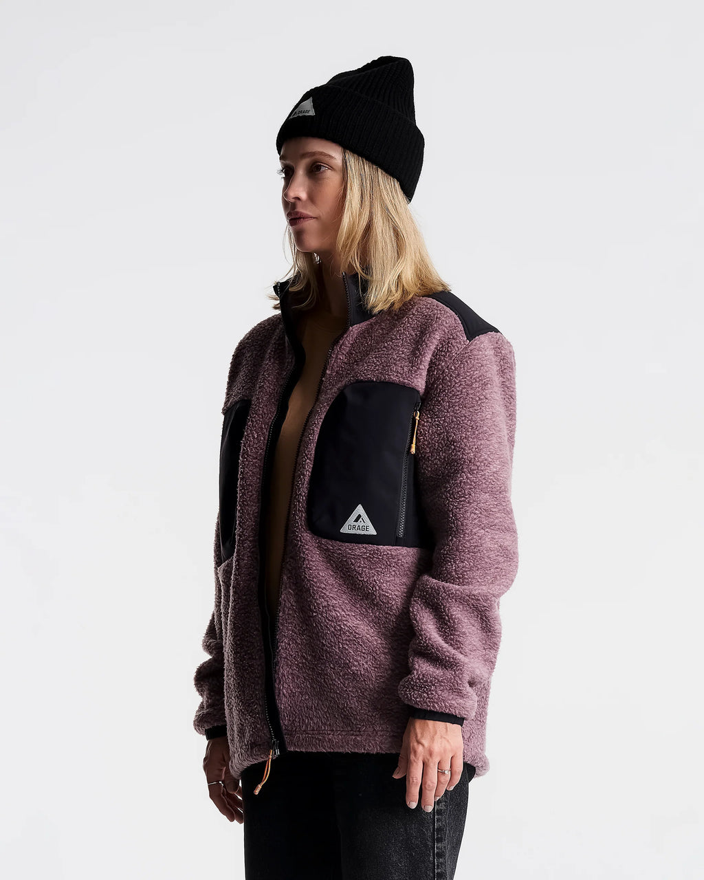 A woman wearing a dusty mauve sherpa fleece jacket with black contrast panels and a black beanie, shown in profile against a light gray studio background.