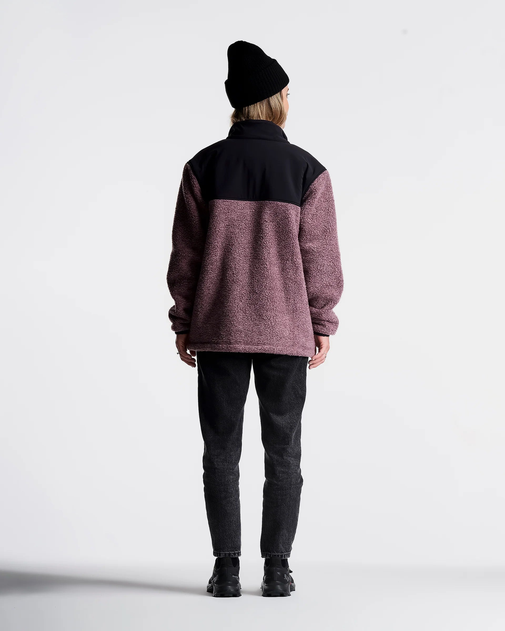 A person shown from the back wearing a color-block fleece jacket with a black upper section and a heathered dusty rose lower section, paired with black jeans and a black beanie, against a plain white studio background.