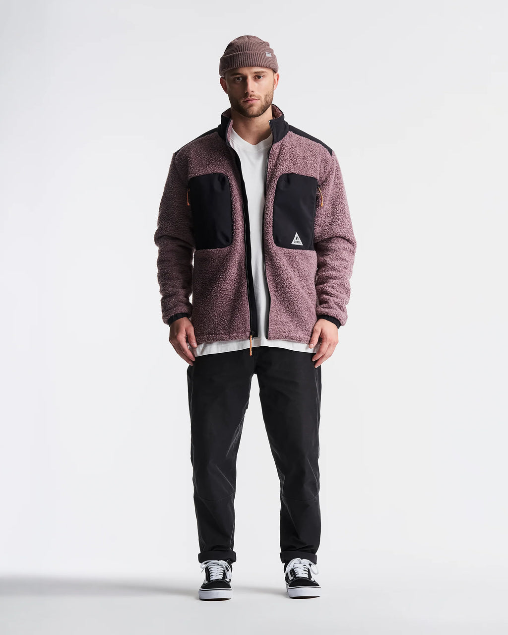 A man wearing a maroon heathered fleece jacket with black contrast panels, a white t-shirt, black pants, and a brown beanie, standing against a light gray studio background.