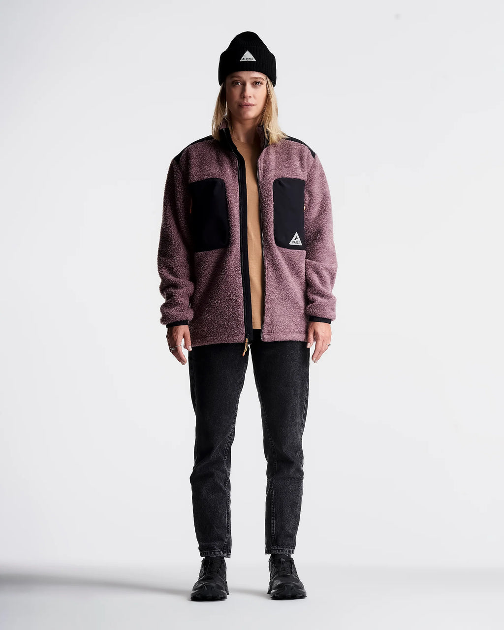A model stands against a light gray studio background wearing a heathered mauve-brown fleece zip-up jacket with black contrast panels and a small white triangular logo, paired with black jeans, black shoes, and a black beanie.