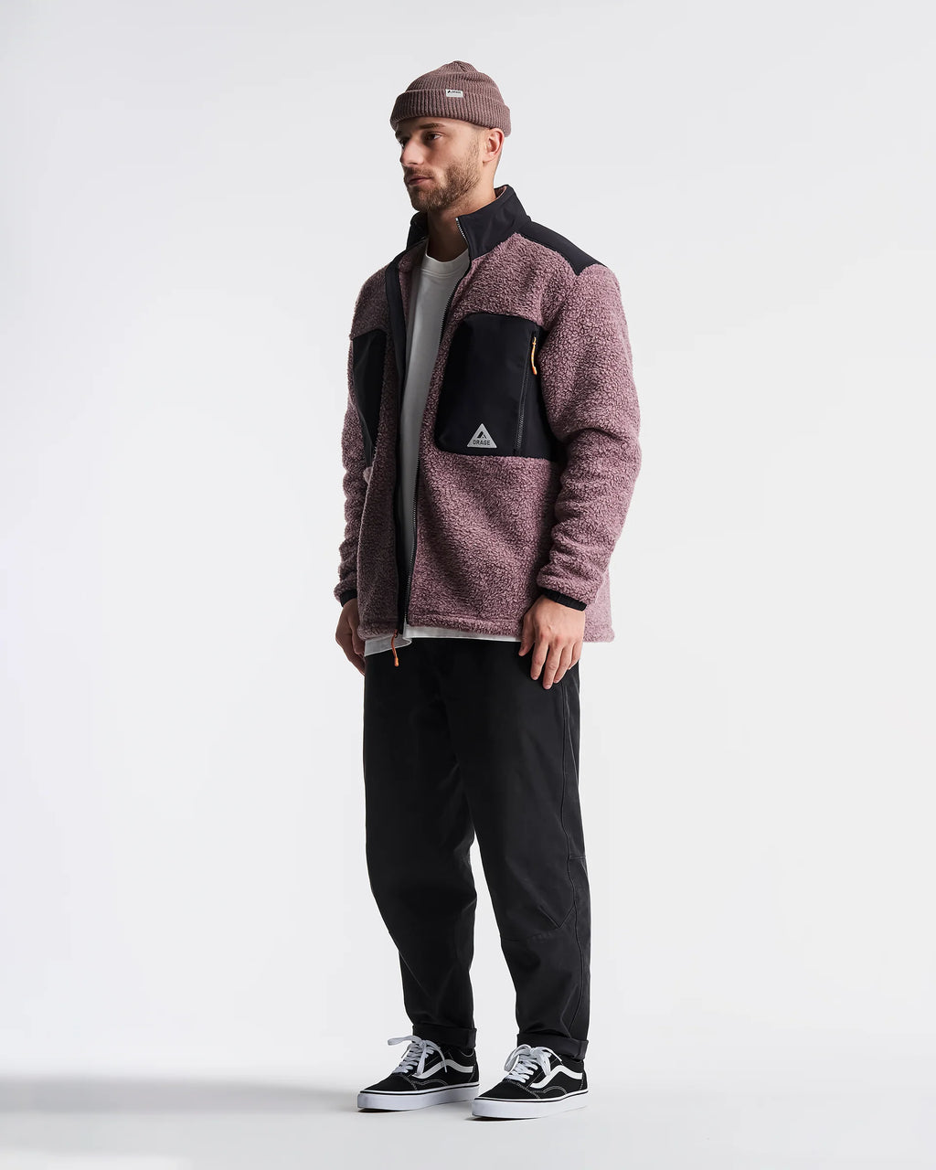 A man wearing a dusty rose and black fleece jacket, black pants, black sneakers, and a brown beanie, standing against a light gray studio background.