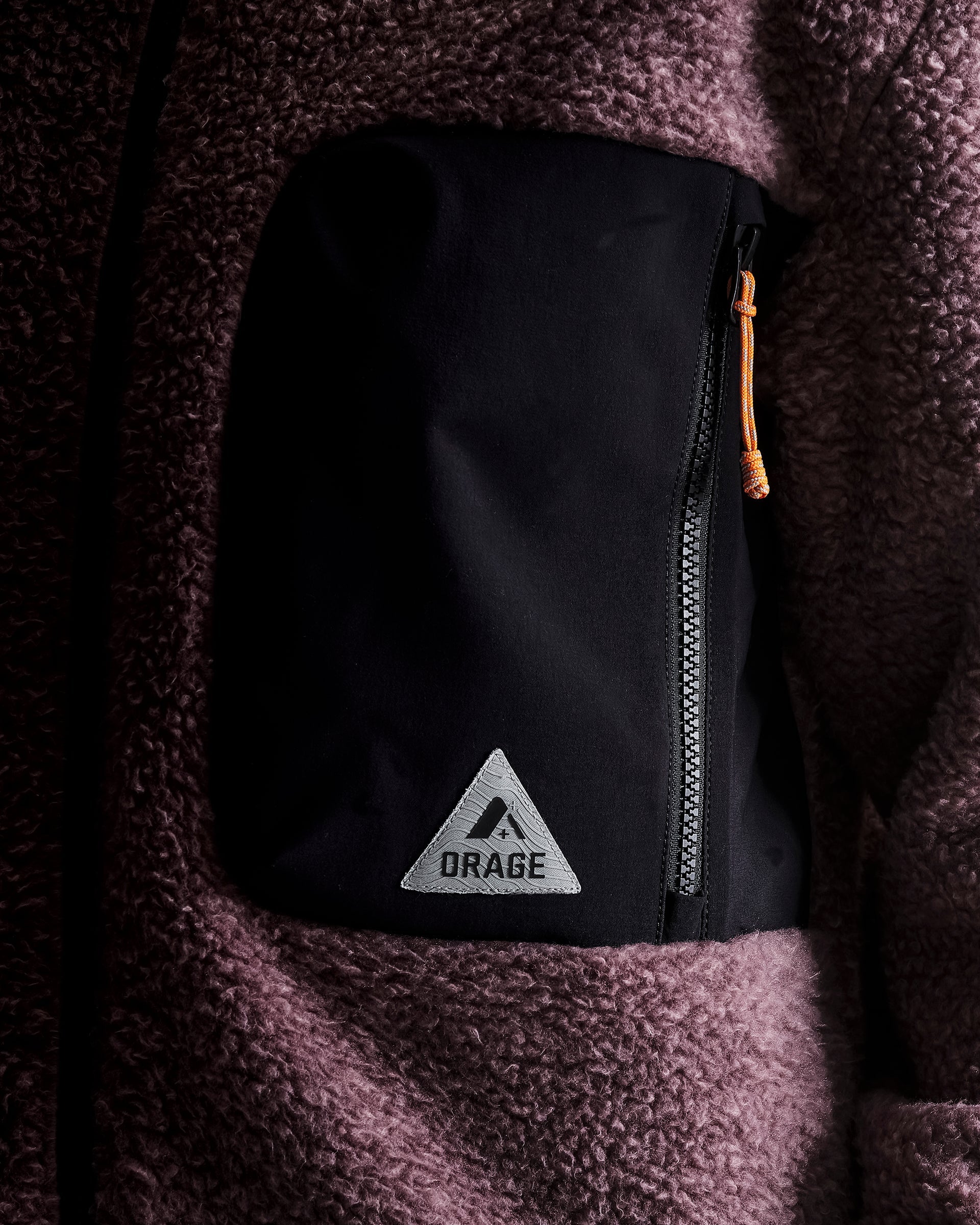 Close-up of a black zippered pouch pocket on a dark brown sherpa fleece jacket, featuring a silver triangular patch with the brand name ORAGE and an orange zipper pull.