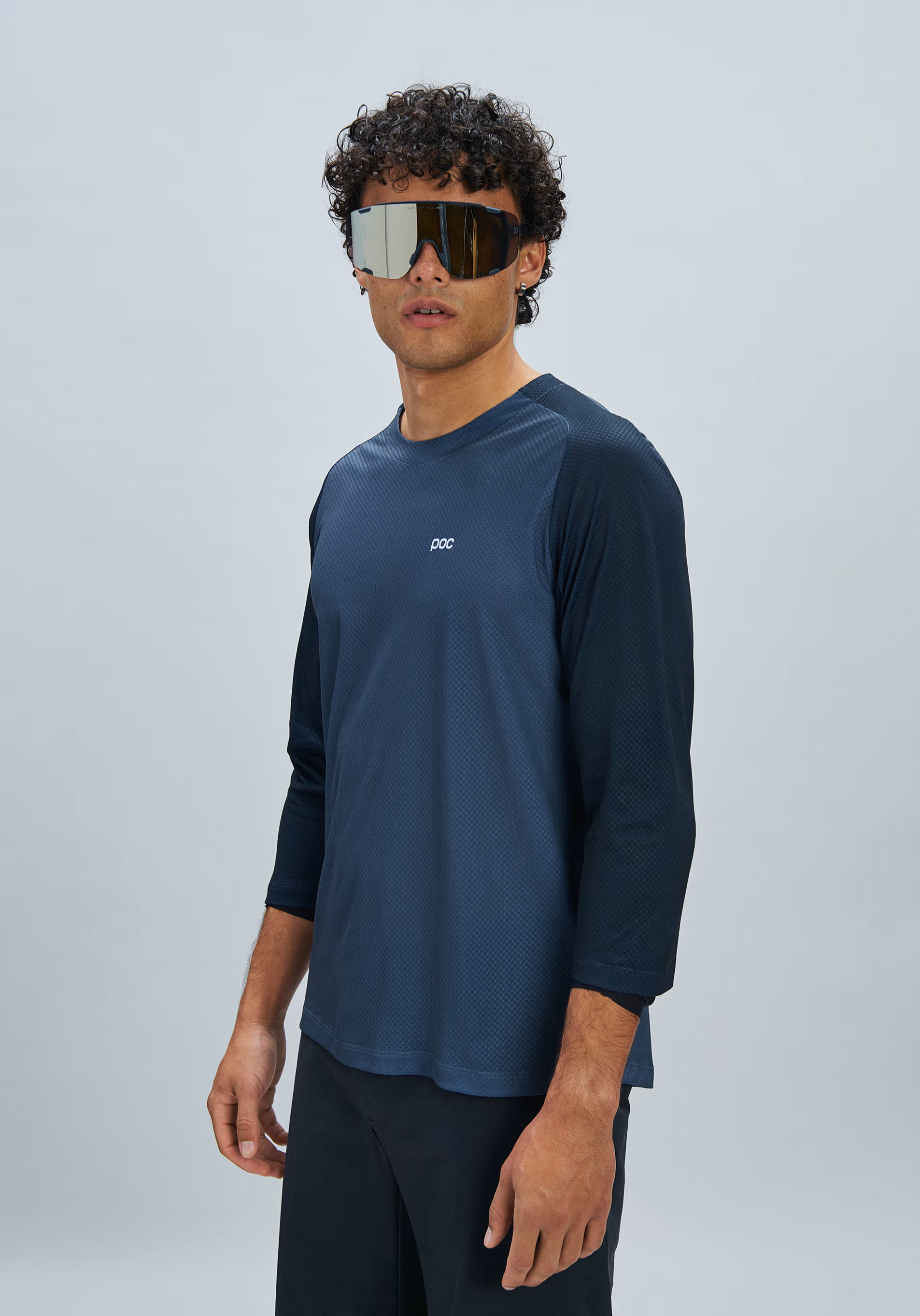 A man wearing a dark blue long-sleeve athletic shirt with three-quarter sleeves and reflective shield sunglasses, standing against a light gray studio background.