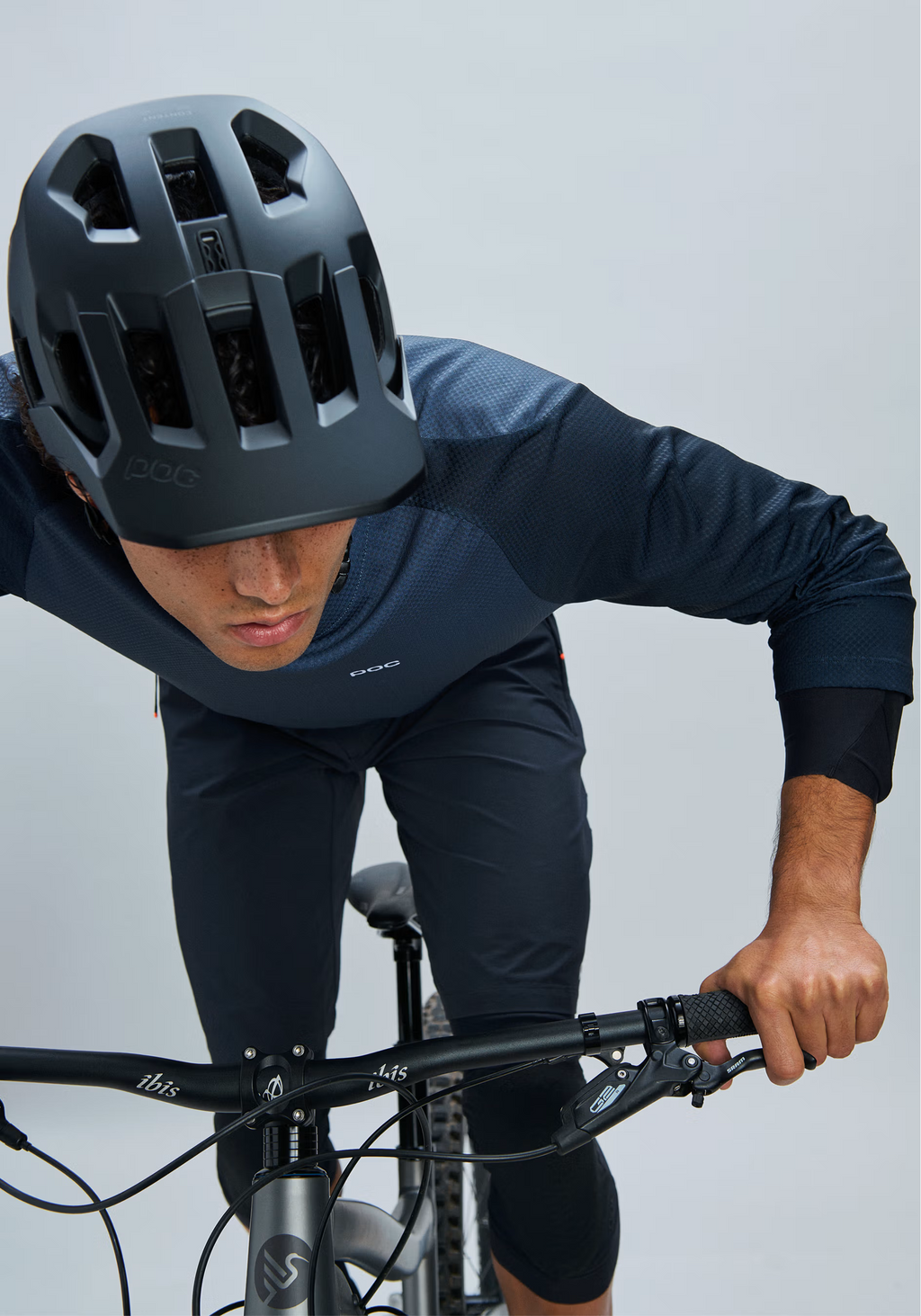 A cyclist wearing a dark gray POC helmet, a navy blue long-sleeve jersey, and matching navy shorts leans forward over a bicycle handlebar against a light gray studio background.