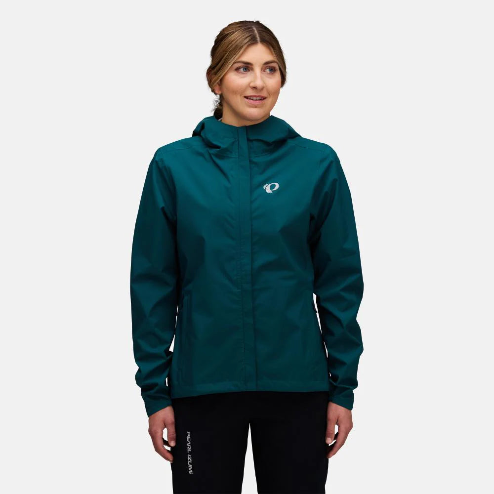 A woman wearing a dark teal hooded jacket with a small white and blue logo on the left chest, paired with black pants, against a light gray studio background.