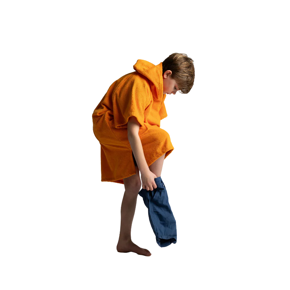 A child wearing an orange hooded towel robe, bending down to remove blue pants against a plain white background.