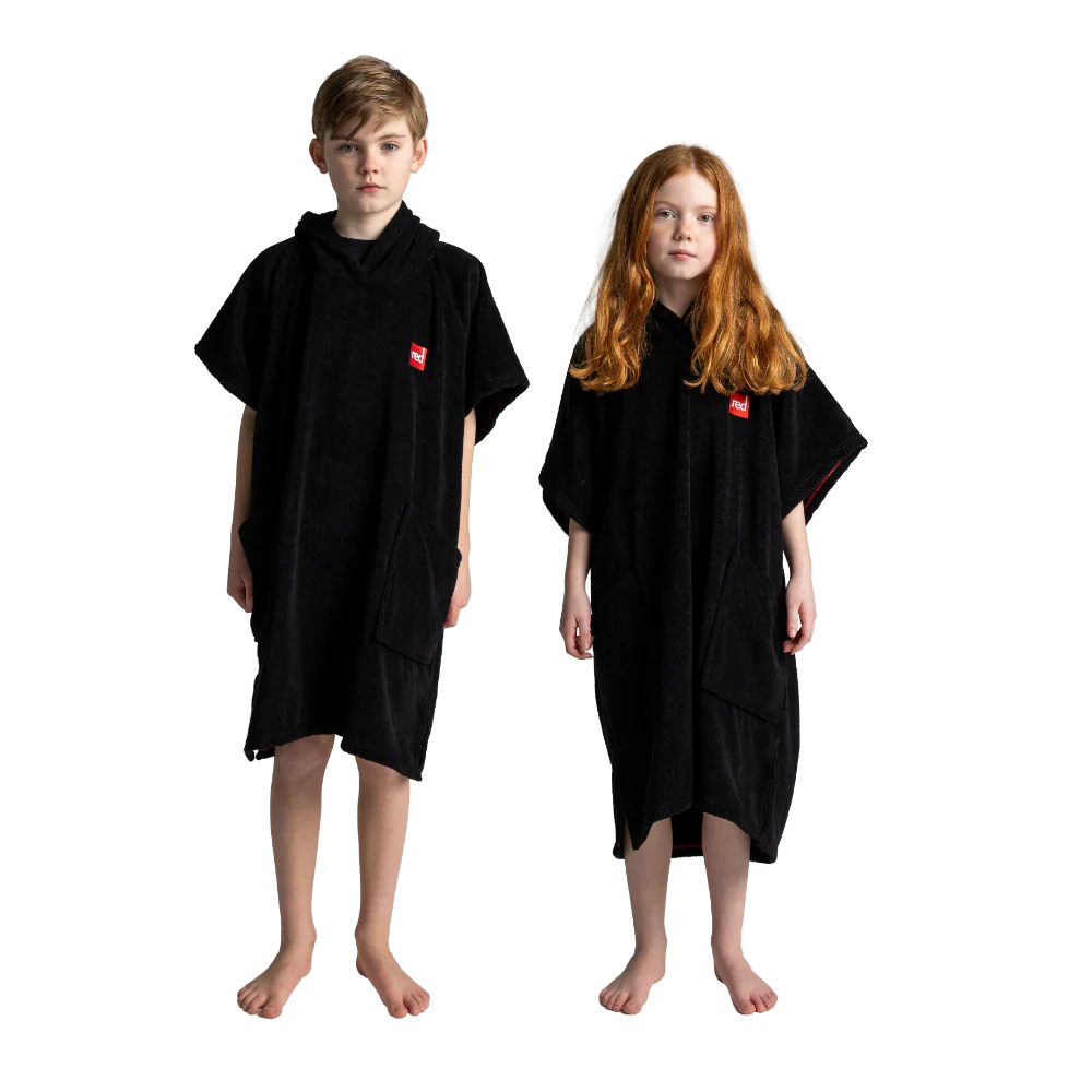 A boy and a girl standing barefoot against a white background, both wearing black hooded towel ponchos with a small red square logo on the chest.