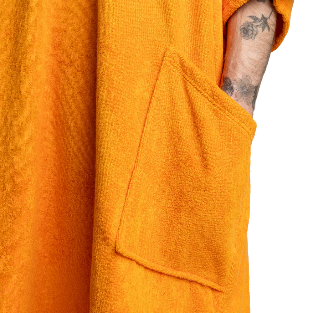 Close-up of a bright orange textured robe or towel with a large front patch pocket and a tattooed arm tucked into the sleeve, against a white background.