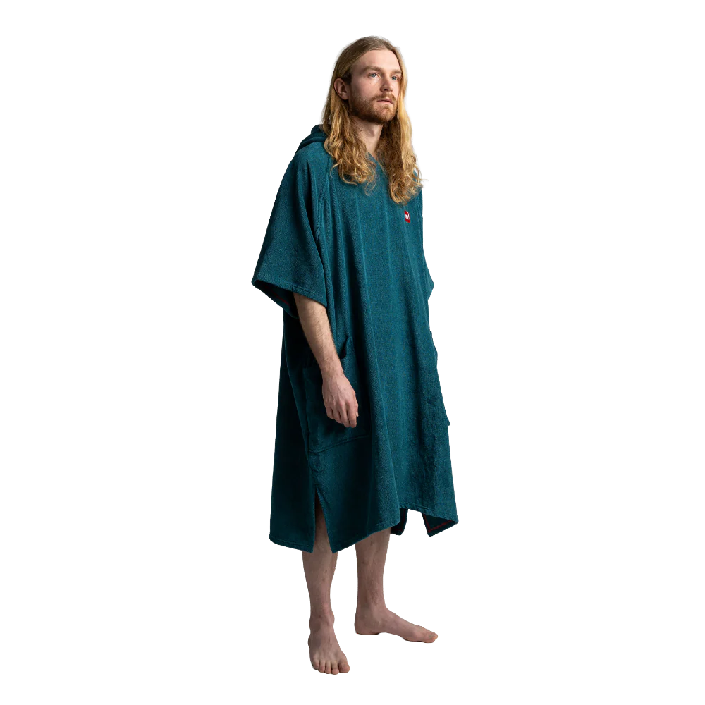 A man with long blonde hair and a beard wearing a dark teal green hooded robe, standing barefoot against a solid white background.