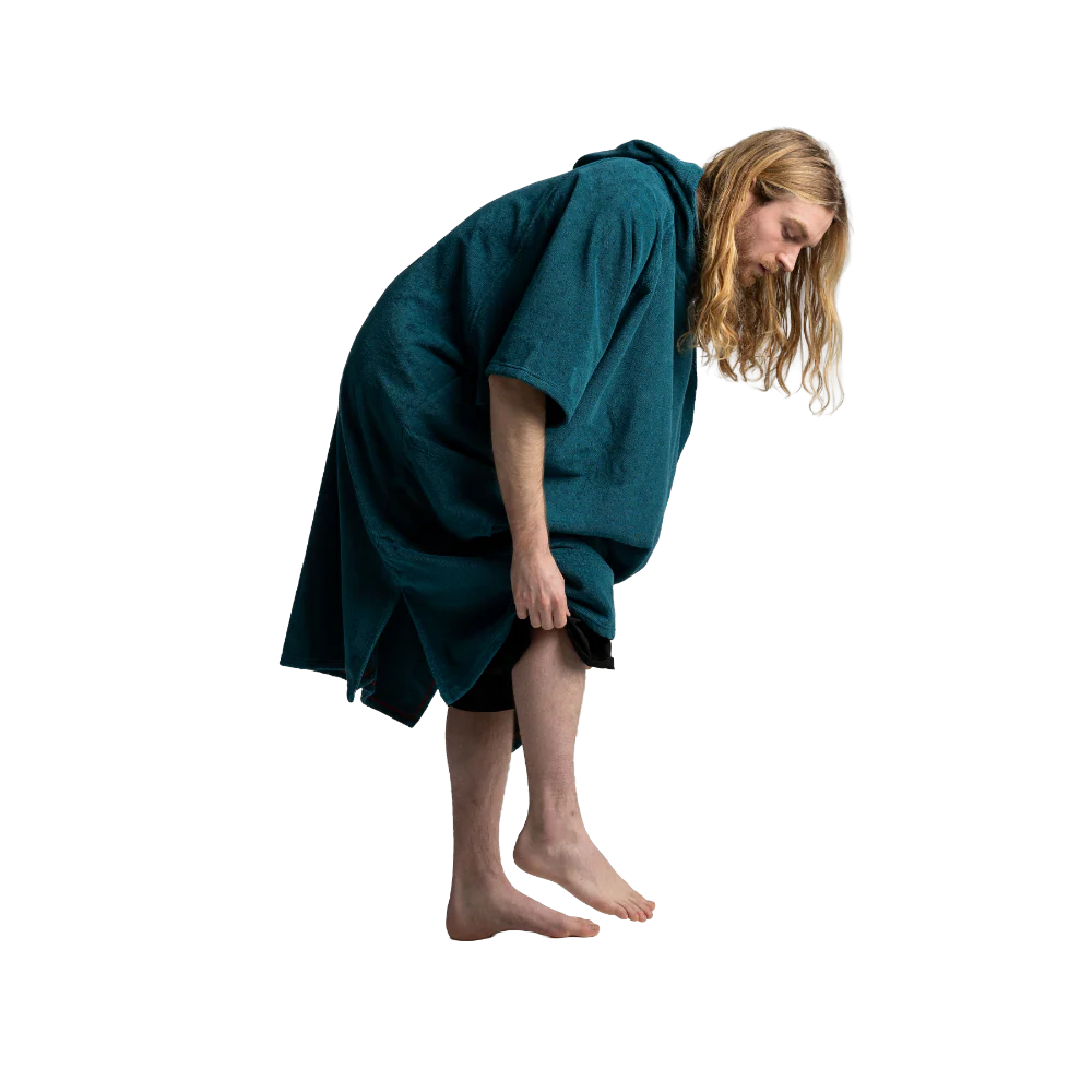 A man with long blonde hair is shown from the waist down, bent forward while wearing a dark teal green hooded robe or poncho over black clothing, against a plain white background.