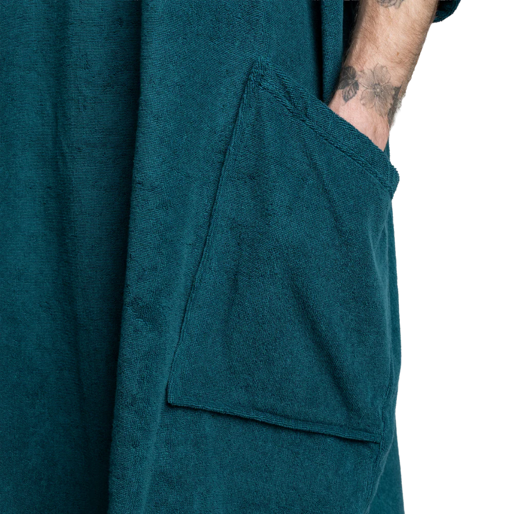 Close-up of a dark teal bathrobe with a large front patch pocket and a person's tattooed arm tucked inside, against a plain white background.