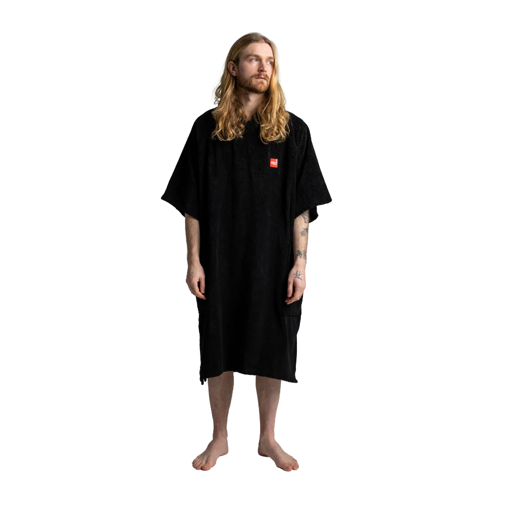 A man with long blonde hair and a beard stands barefoot against a white background, wearing a black short-sleeved robe or poncho-style garment with a small red square logo patch on the left chest.