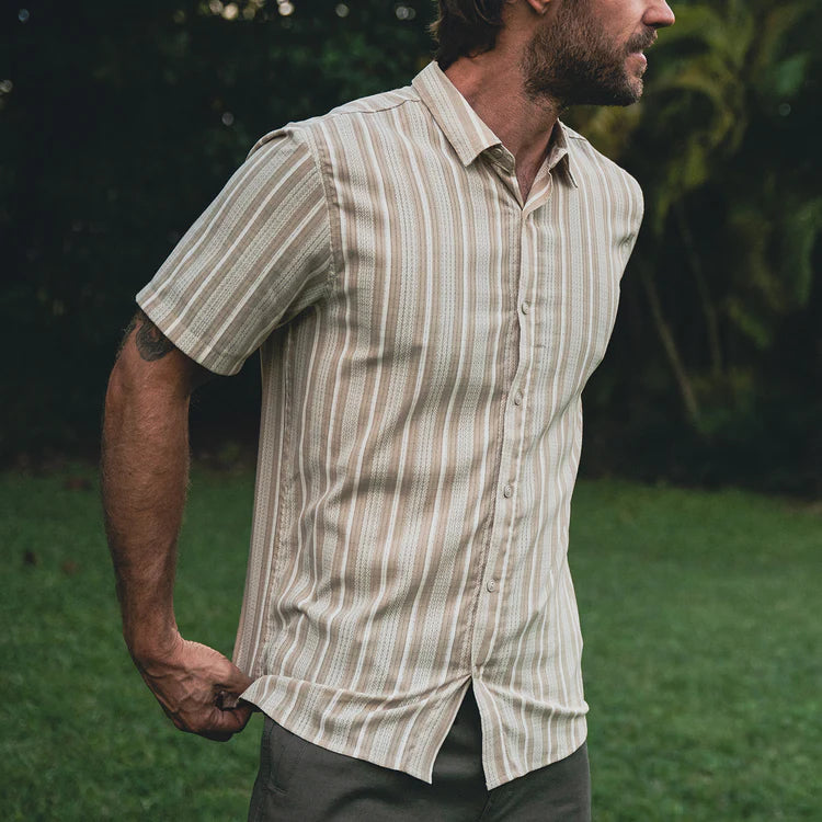 Man wearing a striped shirt standing outdoors with greenery in the background