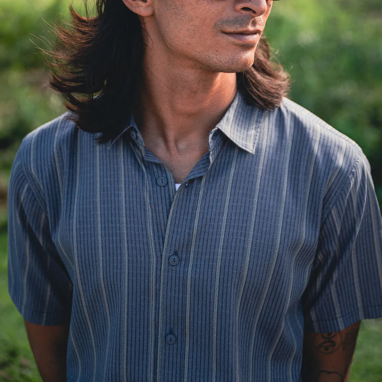 A man wearing a short-sleeve button-up shirt with vertical blue and white stripes, photographed outdoors against a blurred green background.