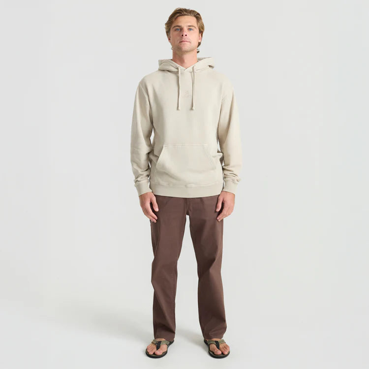 A man modeling a light beige hooded sweatshirt and brown pants against a plain light gray studio background.