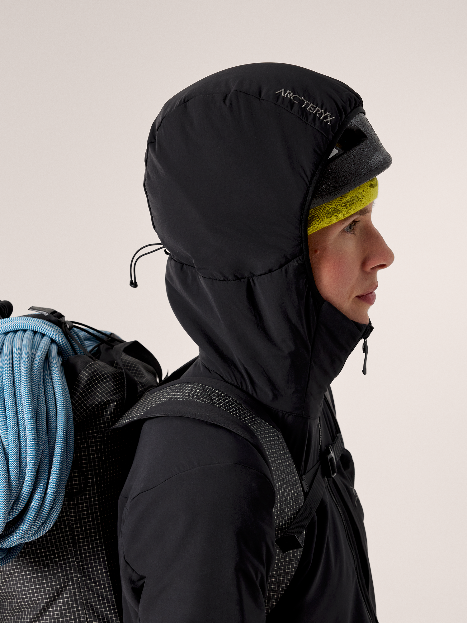 Person wearing a black hooded jacket with a backpack and climbing rope on a plain background