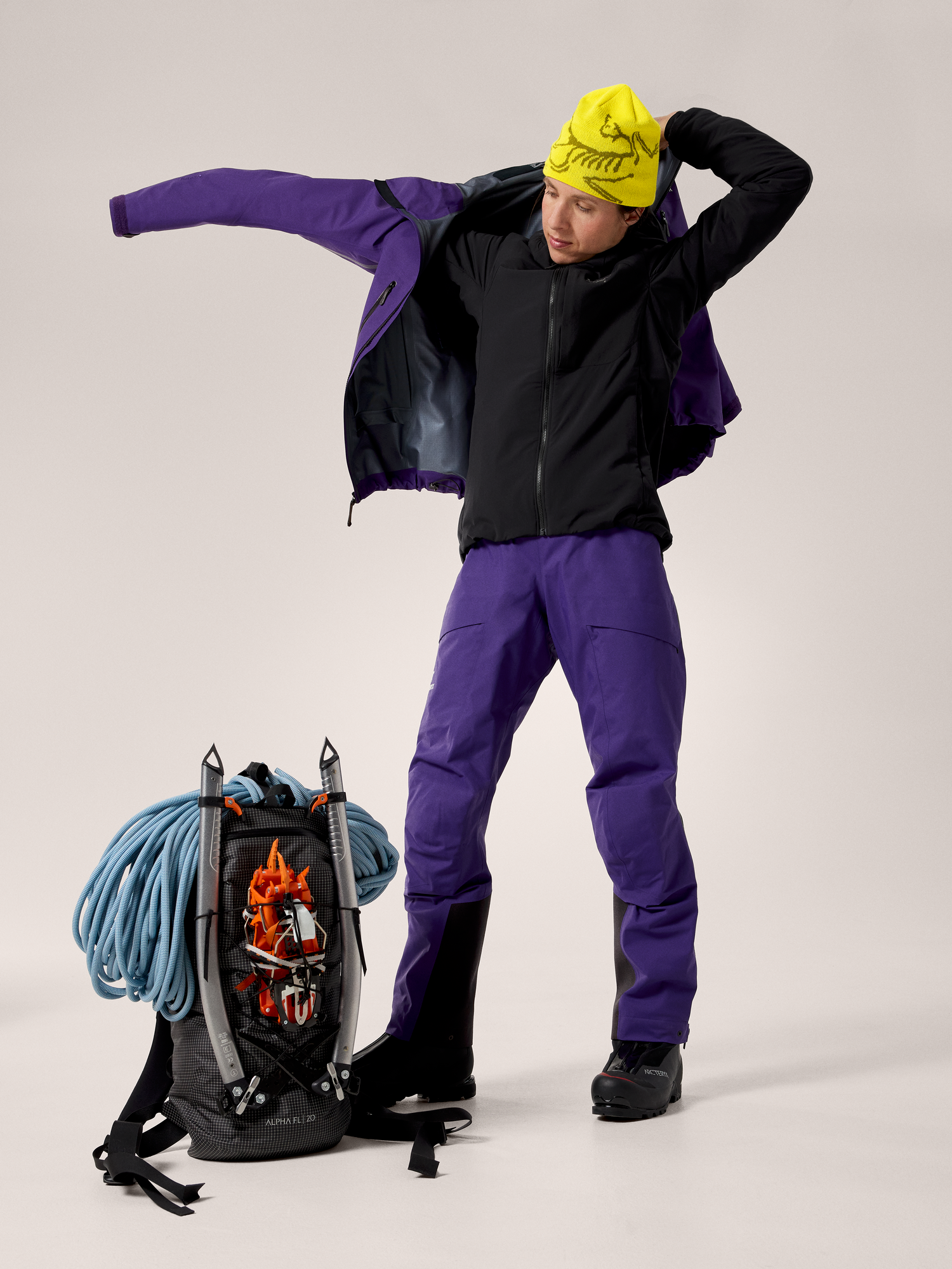 Person wearing a black jacket, purple pants, and a yellow beanie with a backpack and climbing gear on a light gray background