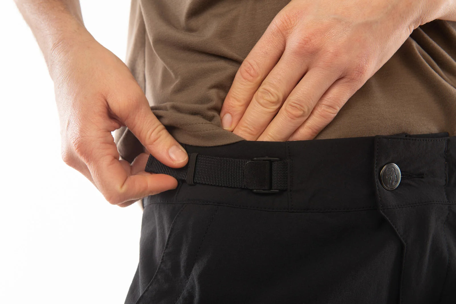 Close-up of a person's hands adjusting a black webbed belt buckle over the waistband of black pants, with a brown shirt visible above.