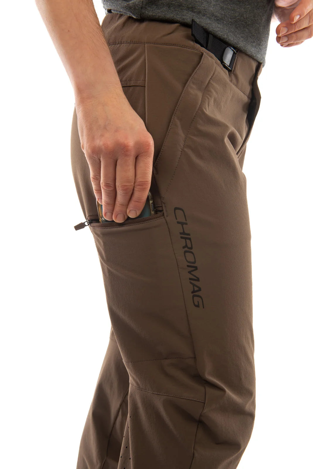 Close-up of a person wearing brown technical outdoor pants with a side zip pocket containing a phone, showing the vertical CHROMAG logo on the thigh.