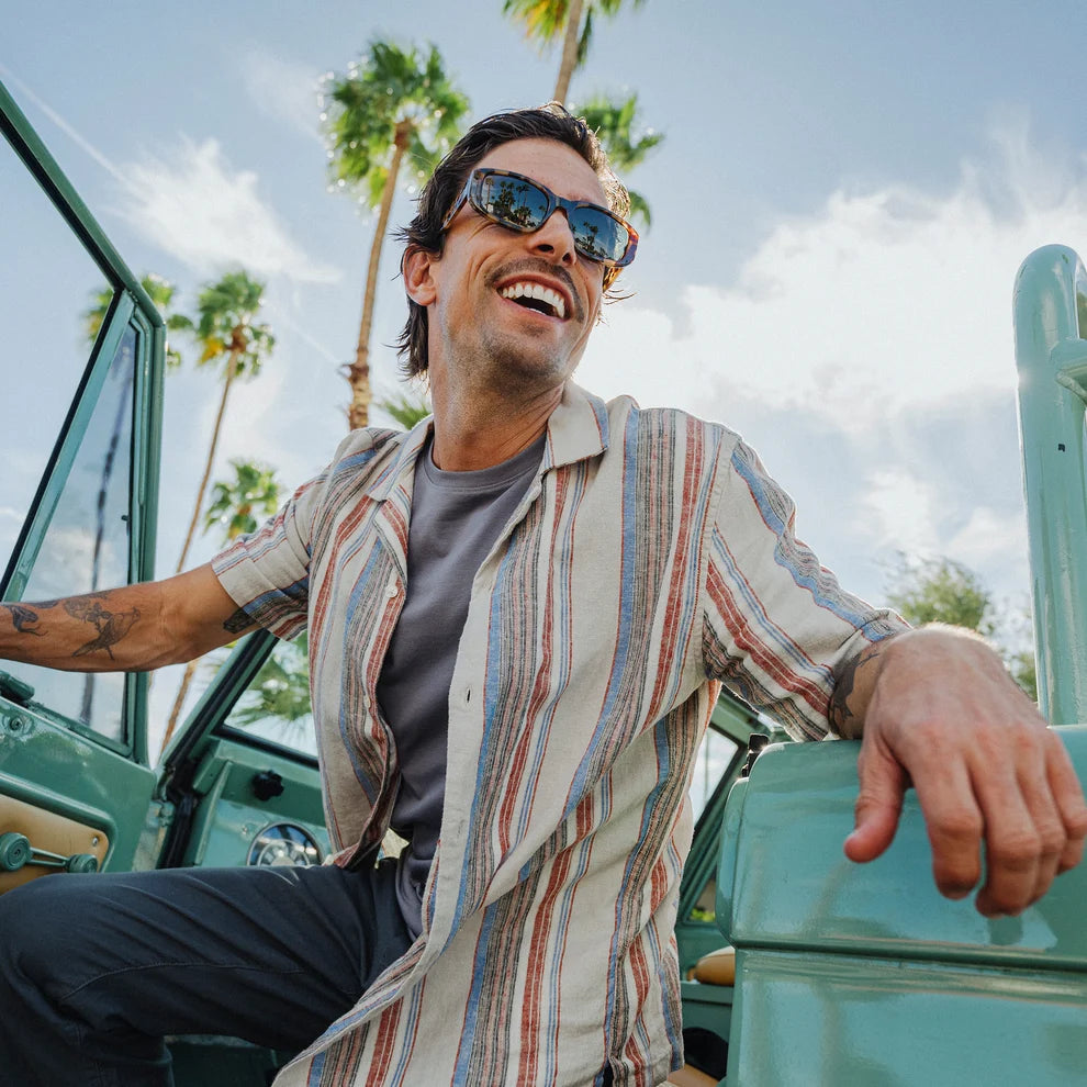 Man wearing sunglasses and a striped shirt sitting in a vintage vehicle with palm trees in the background